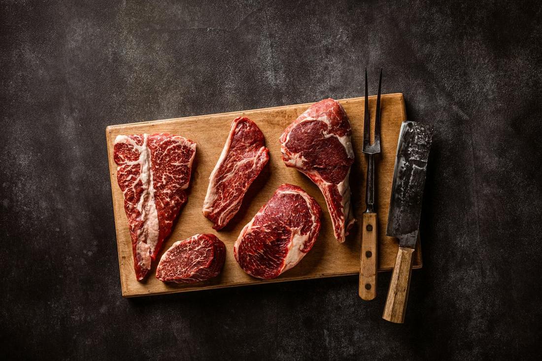 Assorted raw marbled beef steaks on a wooden board with a carving fork and cleaver